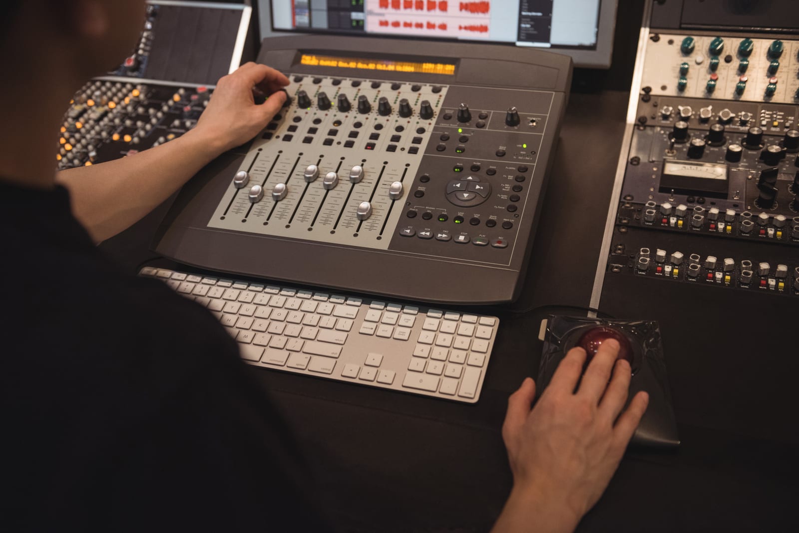 Mãos operam uma superfície de controle e um trackball ao lado de equipamentos de rack.